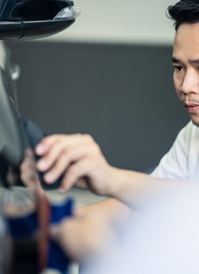 Two Asian car film protection male worker installing film to black color car in garage. Man focus holding film carefully and cutting film in shape. Coating and protective for vehicle business concept.